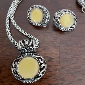 May include: A silver-toned jewelry set featuring a necklace with a pendant and matching earrings. Each piece has a circular, pale yellow center stone surrounded by an ornate, swirling design. The necklace has a braided chain.