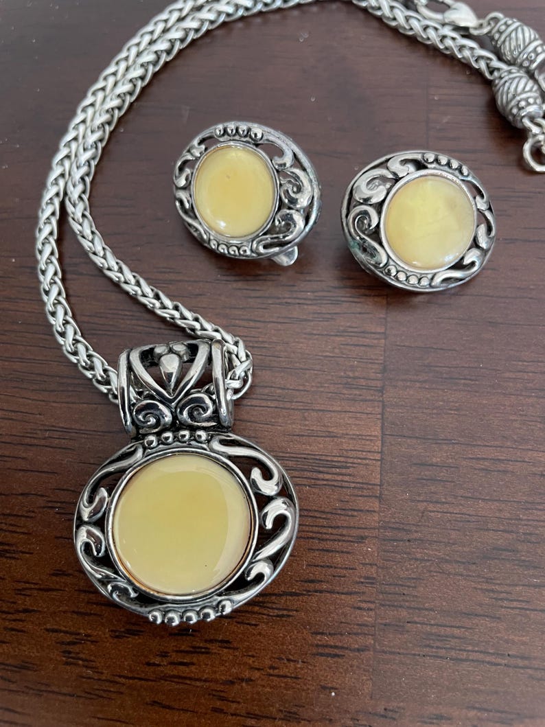 May include: A silver-toned jewelry set featuring a necklace with a pendant and matching earrings. Each piece has a circular, pale yellow center stone surrounded by an ornate, swirling design. The necklace has a braided chain.