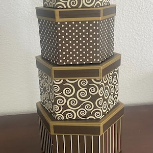 May include: A stack of four hexagonal boxes in varying sizes, featuring a brown and cream color palette. The patterns include cream swirls on brown, white polka dots on brown, and vertical white stripes on a brown base, all with gold trim.
