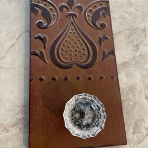 May include: A rectangular metal wall hook featuring a rust-colored finish with an embossed fleur-de-lis pattern and a row of small raised dots. A clear, faceted glass knob is attached to the lower portion of the metal plate.