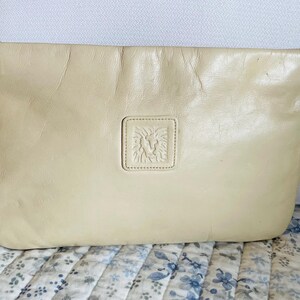 May include: A cream-colored leather clutch with a gold lion head embossed on the front. The clutch has a gold zipper and a gold tag with a lion head on it.