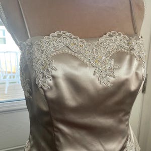 May include: A champagne-colored satin gown with delicate spaghetti straps. The bodice features intricate ivory lace appliqués and beaded embellishments along the neckline and hem. The dress has a fitted silhouette, suitable for formal occasions.