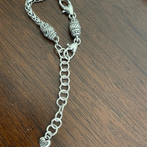 May include: Silver-toned necklace with a braided chain design. The necklace features decorative barrel-shaped beads and heart-shaped clasps. An adjustable chain with a heart-shaped charm completes the design.