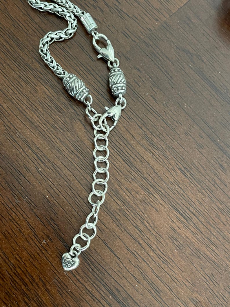 May include: Silver-toned necklace with a braided chain design. The necklace features decorative barrel-shaped beads and heart-shaped clasps. An adjustable chain with a heart-shaped charm completes the design.