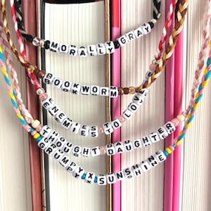 May include: Braided friendship bracelets with white letter beads spelling out phrases like "MORALLY GRAY" and "ENEMIES TO LOVERS." The bracelets are in various colors, including pink, black, and brown, and are displayed against a backdrop of books.