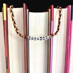 May include: A braided brown and tan bookmark with the word "BOOKWORM" spelled out in white letter beads. The bookmark is draped over the pages of several books with various colored spines, including red, pink, and brown.