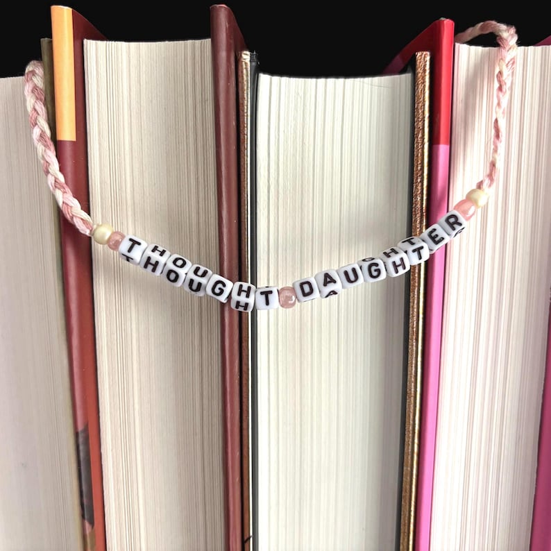 May include: A handmade bookmark with the words "THOUGHT DAUGHTER" spelled out on white letter beads. The bookmark is strung on a pink and white braided cord, and is placed between the pages of several books.