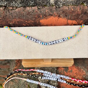 May include: Handmade friendship bracelets with colorful braided cords and white letter beads. The top bracelet reads "GRUMPY X SUNSHINE". Other bracelets below read "ENEMIES TO LOVERS", "BOOKWORM", "MORALLY GRAY", and "DAUGHTER".