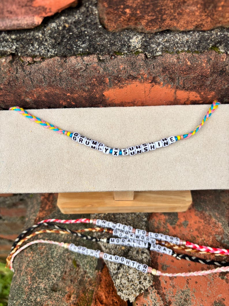 May include: Handmade friendship bracelets with colorful braided cords and white letter beads. The top bracelet reads "GRUMPY X SUNSHINE". Other bracelets below read "ENEMIES TO LOVERS", "BOOKWORM", "MORALLY GRAY", and "DAUGHTER".