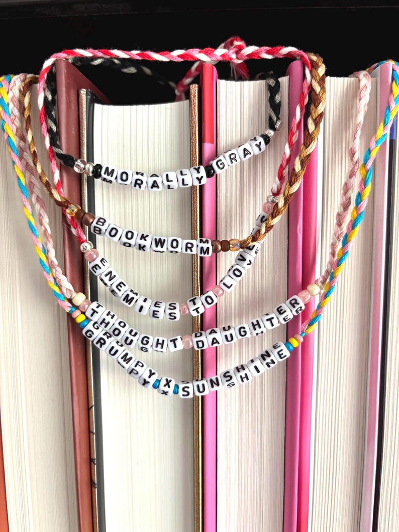 May include: Braided friendship bracelets with white letter beads spelling out phrases like "MORALLY GRAY" and "ENEMIES TO LOVERS." The bracelets are in various colors, including pink, black, and brown, and are displayed against a backdrop of books.
