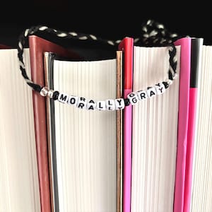 May include: A handmade beaded bookmark with the words "MORALLY GRAY" spelled out in black letters on white beads. The bookmark has a black and white braided cord and is placed between the pages of several books.