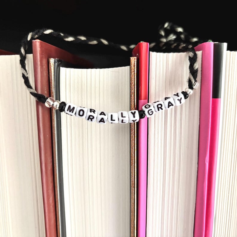 May include: A handmade beaded bookmark with the words "MORALLY GRAY" spelled out in black letters on white beads. The bookmark has a black and white braided cord and is placed between the pages of several books.