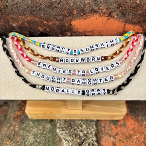 May include: A display of beaded bracelets with various phrases spelled out in white letter beads. The phrases include "GRUMPY X SUNSHINE", "BOOKWORM", "ENEMIES TO LOVERS", "THOUGHT DAUGHTER", and "MORALLY GRAY". The bracelets are in different colors.
