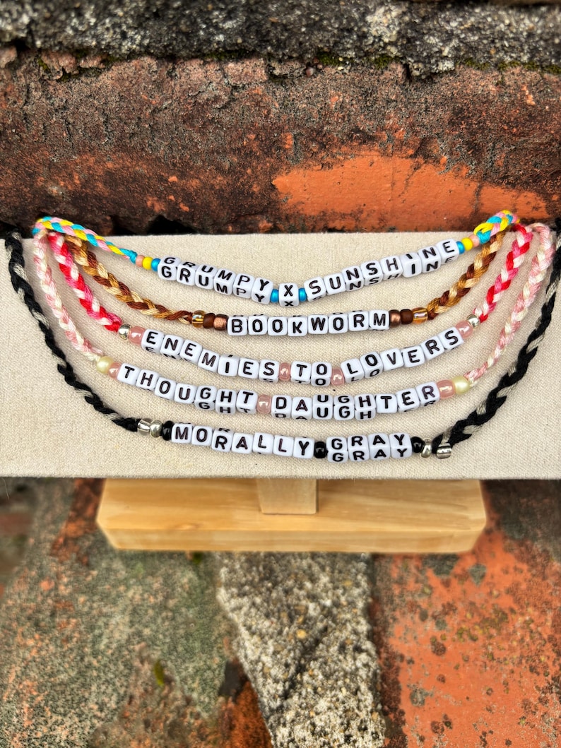 May include: A display of beaded bracelets with various phrases spelled out in white letter beads. The phrases include "GRUMPY X SUNSHINE", "BOOKWORM", "ENEMIES TO LOVERS", "THOUGHT DAUGHTER", and "MORALLY GRAY". The bracelets are in different colors.
