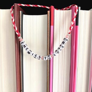 May include: A handmade bookmark with the words "ENEMIES TO LOVERS" spelled out on white letter beads. The beads are strung on a red and white braided cord. The bookmark is placed between the pages of several books.