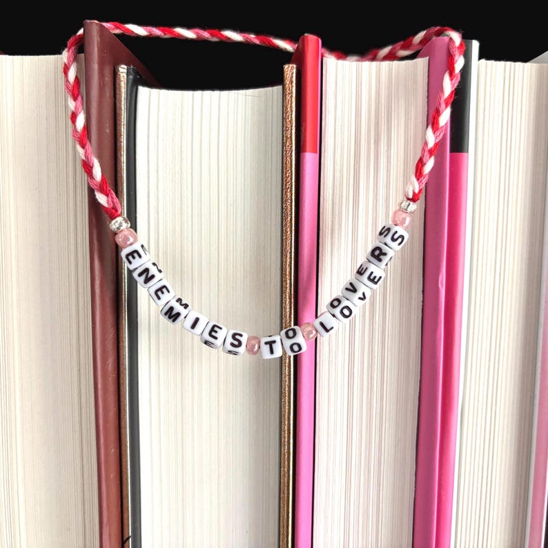 May include: A handmade bookmark with the words "ENEMIES TO LOVERS" spelled out on white letter beads. The beads are strung on a red and white braided cord. The bookmark is placed between the pages of several books.