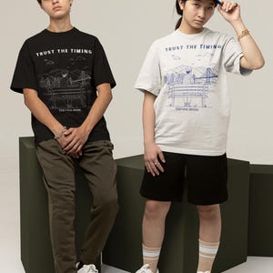 Puede incluir: Dos personas con camisetas a juego con el texto "TRUST THE TIMING" y un dibujo lineal de un paisaje urbano. Una camiseta es negra, la otra gris claro. Ambos llevan zapatillas blancas. Una persona lleva una gorra azul.