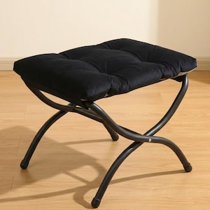 May include: A black upholstered ottoman with a tufted cushion. The ottoman has a curved, dark metal frame. The legs are curved and meet in the middle. The ottoman is designed for seating.