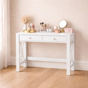 May include: A white vanity table with two drawers and silver knobs. The table has an X-shaped support on each side. Various beauty products, a mirror, and a small pink box are arranged on top, creating a clean and organized aesthetic.