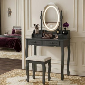 May include: A dark gray vanity table with an oval mirror featuring a built-in light. The vanity has two drawers, a stool with a cream-colored seat, and decorative accents. Makeup brushes and a palette are visible on the table.