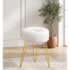 May include: A white and gold patterned ottoman with gold hairpin legs. The round seat features a white background with gold vein-like designs. The gold legs are thin and angled, providing a modern touch. A decorative piece for any living space.