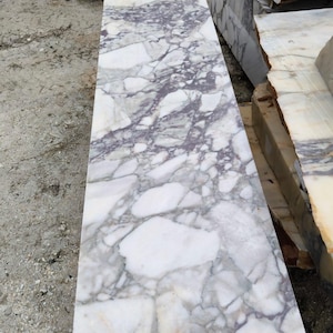 May include: A rectangular slab of marble with a white and purple veined pattern. The stone has a polished surface and is likely intended for use in construction or design projects. Other marble slabs are visible in the background.