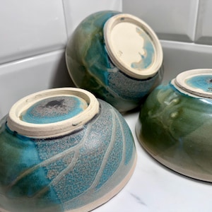 May include: Three ceramic bowls with a glossy finish. The bowls are glazed in shades of teal, green, and blue, with a speckled pattern on the exterior. The bottoms are white with a teal center. The bowls are arranged on a white surface.