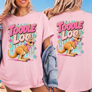 Op de afbeelding: Lichtroze T-shirt met een cartoon kangoeroe met een zonnebril en een roze bandana. De tekst "Toodle Loo" staat boven de kangoeroe in roze en witte letters, met sterren en strepen op de achtergrond.
