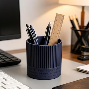 May include: A navy blue pen holder with vertical ribbed detailing, holding pens and a wooden ruler. The pen holder has the quote "You don't have to see the whole staircase. Just take the next step" adapted from Martin Luther King Jr.
