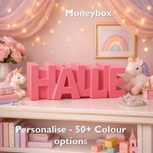 May include: A pink moneybox spelling "HALLE" is displayed on a white shelf. The word "Moneybox" is at the top. The text "Personalise - 50+ Colour options" is at the bottom. The scene includes a unicorn, a crown, and a rainbow picture.