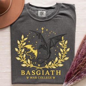 May include: Dark gray t-shirt featuring a black dragon design with a gold dragon and laurel wreath. The text "BASGIATH" and "WAR COLLEGE" are printed in gold. The shirt is made by Comfort Colors.