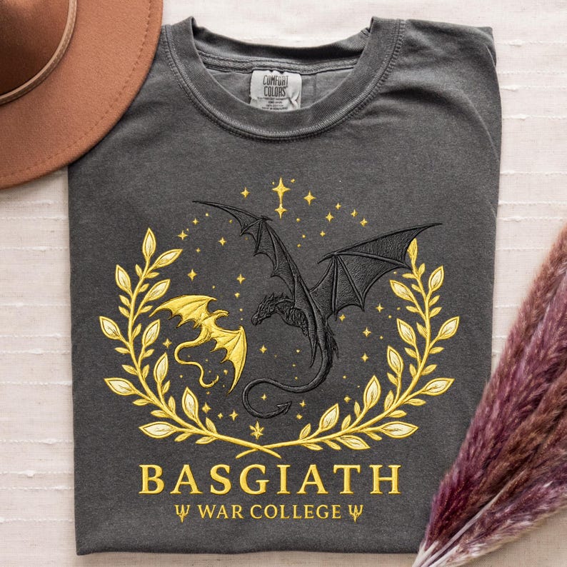 May include: Dark gray t-shirt featuring a black dragon design with a gold dragon and laurel wreath. The text "BASGIATH" and "WAR COLLEGE" are printed in gold. The shirt is made by Comfort Colors.