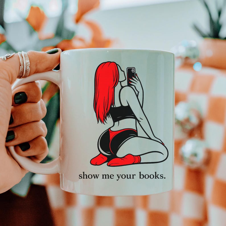 Pu&ograve; includere: Tazza in ceramica bianca con grafica di una persona con capelli rossi che si fa un selfie, con top nero e pantaloncini rossi. Il testo "show me your books" &egrave; stampato sotto. La tazza ha un manico bianco.