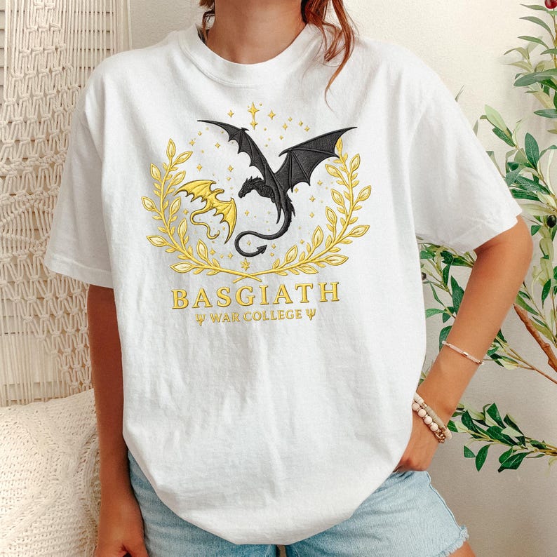 May include: White t-shirt featuring a gold and black dragon design within a gold laurel wreath. The text "BASGIATH WAR COLLEGE" is printed below the dragon illustration. The shirt is a casual, short-sleeved style.