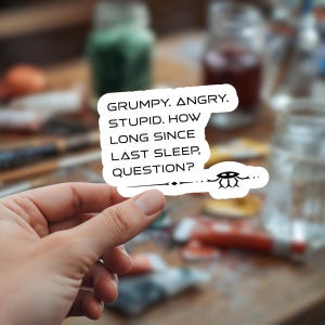 May include: A white sticker with black text that reads "GRUMPY. ANGRY. STUPID. HOW LONG SINCE LAST SLEEP, QUESTION?" The sticker has a small black alien-like graphic and is held by a hand.