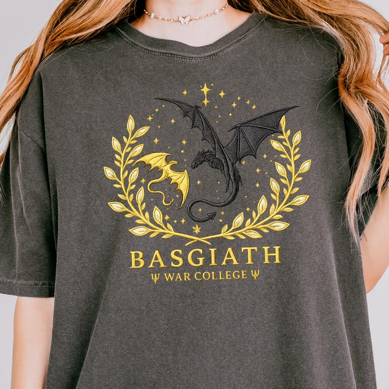 May include: Dark gray t-shirt featuring a gold and black dragon design within a laurel wreath. The text "BASGIATH" and "WAR COLLEGE" are printed below the design. The shirt is made of a soft material.