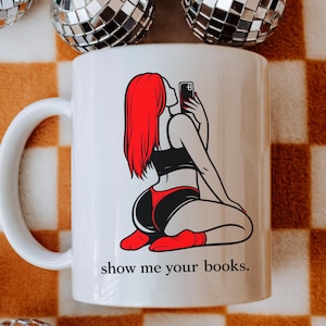Pu&ograve; includere: Tazza in ceramica bianca con un'illustrazione a cartoni animati di una persona con i capelli rossi che si fa un selfie. La persona indossa lingerie nera e rossa. Il testo "show me your books" &egrave; stampato sotto.