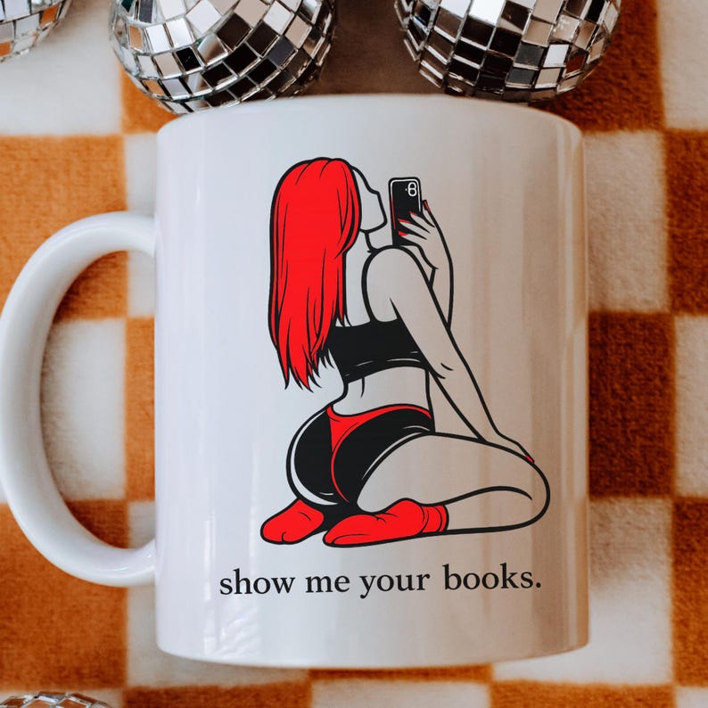 Pu&ograve; includere: Tazza in ceramica bianca con un'illustrazione a cartoni animati di una persona con i capelli rossi che si fa un selfie. La persona indossa lingerie nera e rossa. Il testo "show me your books" &egrave; stampato sotto.