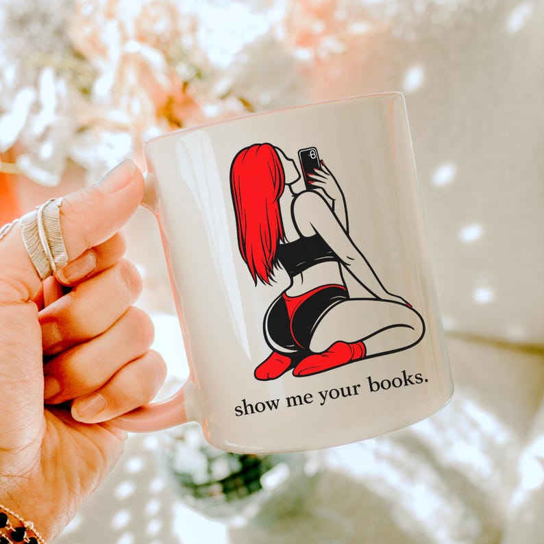 Pu&ograve; includere: Tazza in ceramica bianca con la grafica di una persona con i capelli rossi che si fa un selfie. La persona indossa lingerie nera e rossa. La tazza ha il testo "show me your books."