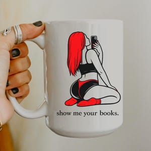 Pu&ograve; includere: Tazza in ceramica bianca con la grafica di una donna con i capelli rossi che si fa un selfie. Indossa un top nero, pantaloncini rossi e neri e calzini rossi. La tazza ha il testo "show me your books."