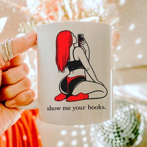 Pu&ograve; includere: Tazza in ceramica bianca con la grafica di una persona con i capelli rossi che si fa un selfie. La persona indossa lingerie nera e rossa. Il testo "show me your books" &egrave; stampato sotto.