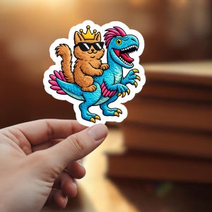 Könnte beinhalten: Ein Aufkleber mit einer Cartoon-Katze mit Sonnenbrille und Krone, die auf einem blauen Dinosaurier reitet. Die Katze ist orange, der Dinosaurier hat einen rosa Kamm und gelbe Krallen. Der Aufkleber hat einen weißen Rand.