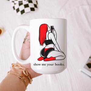 Pu&ograve; includere: Tazza in ceramica bianca con una grafica di una persona con i capelli rossi che si fa un selfie, che indossa abiti neri e rossi. Il testo "show me your books" &egrave; stampato sotto. La tazza ha un manico grande.