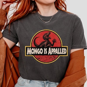 Può includere: T-shirt grigio scuro con grafica di dinosauro all'interno di un cerchio rosso e giallo e la scritta "MONGO IS APPALLED" in un rettangolo. Il dinosauro è nero e rosso.