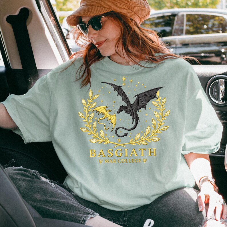 May include: A light green t-shirt featuring a black dragon with outstretched wings and a yellow dragon, both within a gold laurel wreath. The text "BASGIATH" and "WAR COLLEGE" are printed below the dragons. The shirt is a casual wear item.