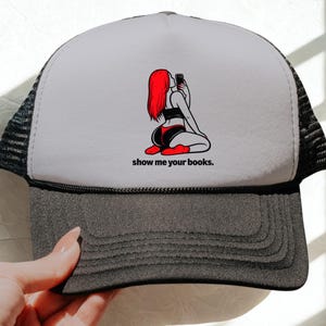 Może przedstawiać: Czapka typu trucker z szarym daszkiem i białym panelem przednim. Na panelu przednim znajduje się grafika przedstawiająca osobę z rudymi włosami robiącą selfie, z napisem "show me your books."