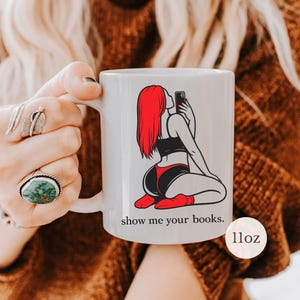 Pu&ograve; includere: Tazza in ceramica bianca con la grafica di una persona con i capelli rossi che si fa un selfie. La persona indossa un top nero, pantaloncini rossi e neri e calzini rossi. La tazza ha il testo "show me your books." ed &egrave; di 312 grammi.