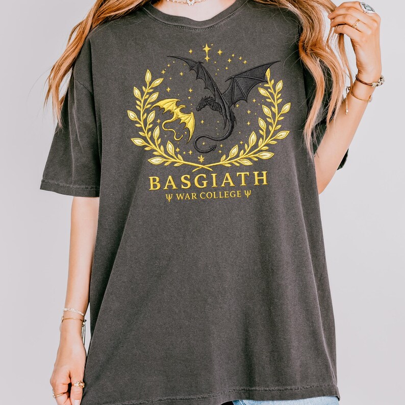 May include: Dark gray t-shirt featuring a dragon design within a golden laurel wreath. The design includes a black dragon and a yellow dragon, with the word "BASGIATH" and "WAR COLLEGE" printed in gold.