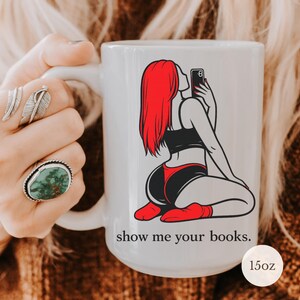 Pu&ograve; includere: Tazza in ceramica bianca con grafica di una donna dai capelli rossi che si fa un selfie, vestita di nero e rosso. La tazza ha il testo "show me your books." ed &egrave; da 15oz.