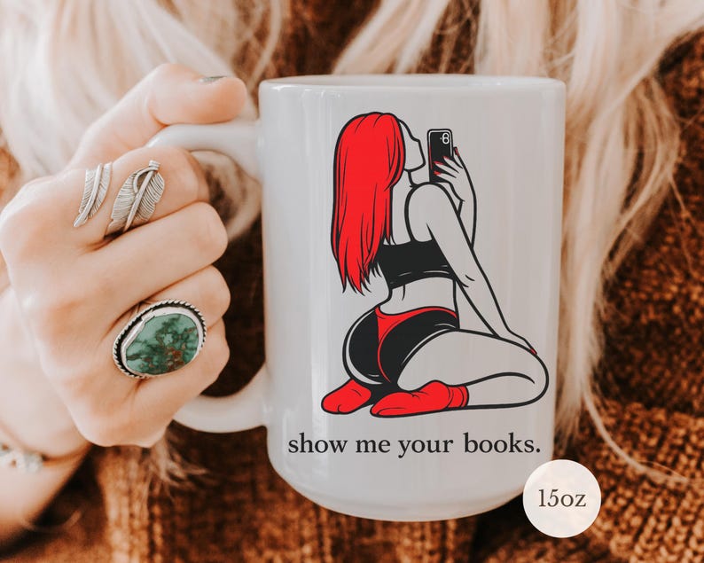 Pu&ograve; includere: Tazza in ceramica bianca con grafica di una donna dai capelli rossi che si fa un selfie, vestita di nero e rosso. La tazza ha il testo "show me your books." ed &egrave; da 15oz.